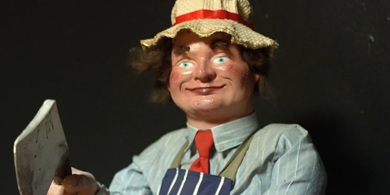 old plastic doll holding an axe. Wearing white hat with red ribbon, a red rie, light blue striped shirt and dark blue stripe gown